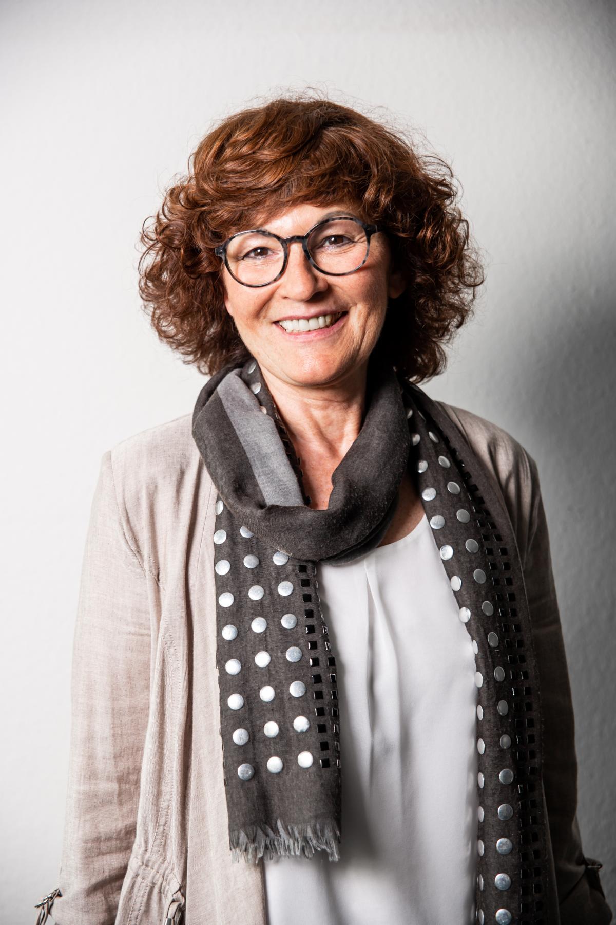 Frau mit lockigem Haar, trägt hellen Cardigan und gepunkteten Schal, lächelt leicht.