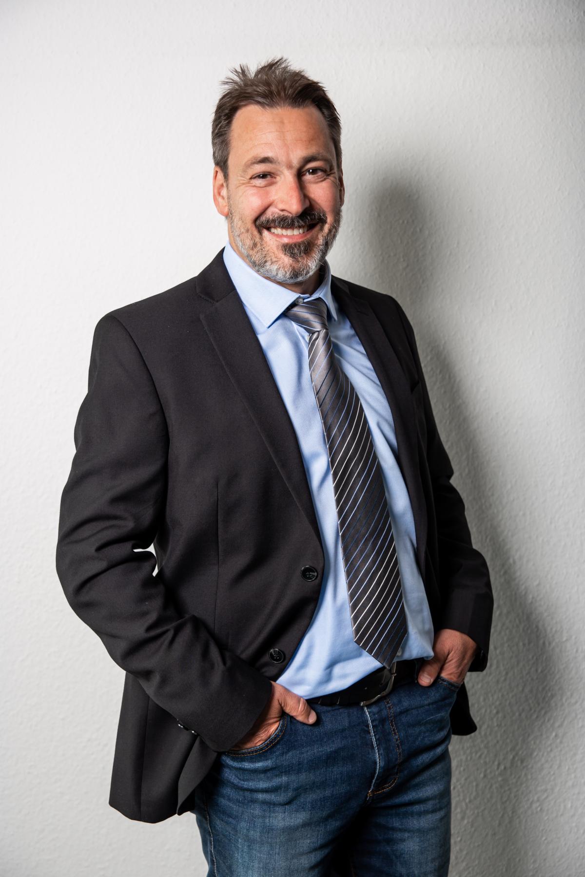 Mann mit Bart, dunklem Sakko und gestreifter Krawatte.